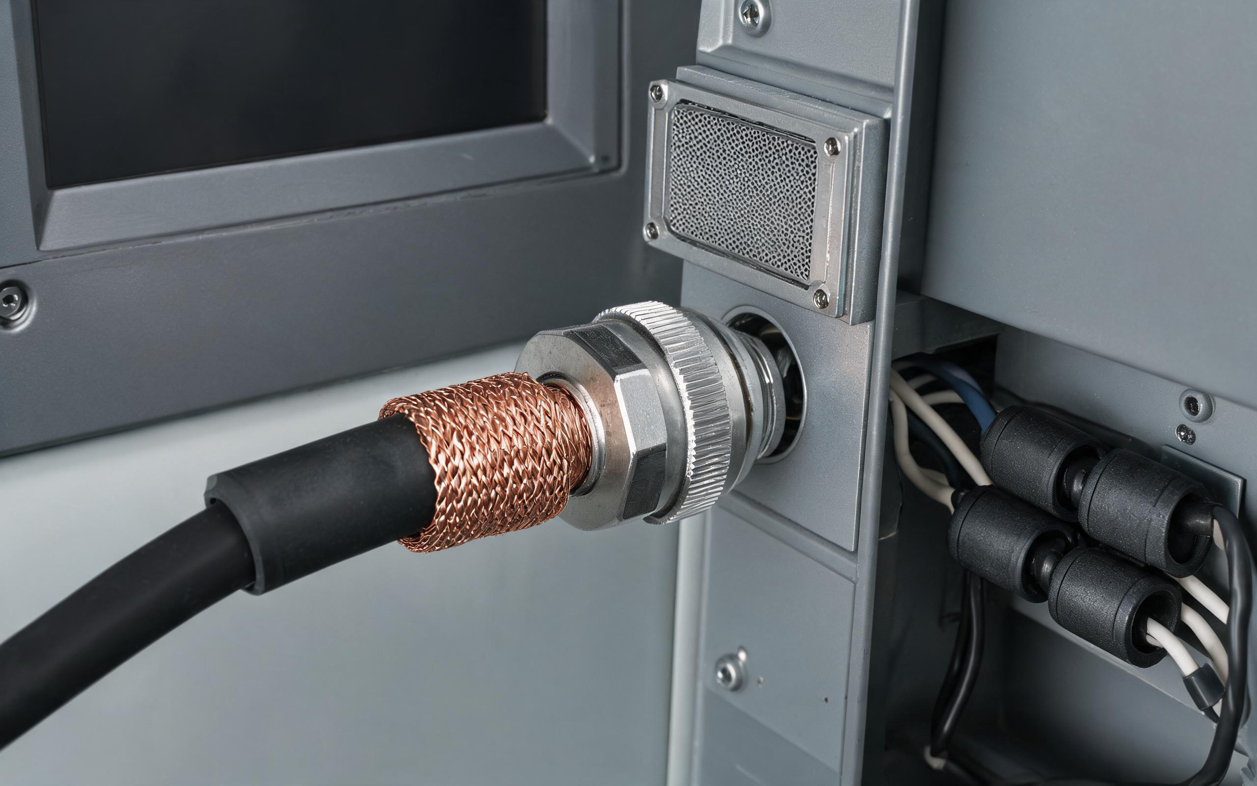 EMC cable gland with 360 degree shield termination on a shielded cable entering the metal enclosure of panel-mounted industrial display monitors, clamp-on ferrite cores on internal harness and a panel filter, realistic industrial engineering photography, no readable text, no people